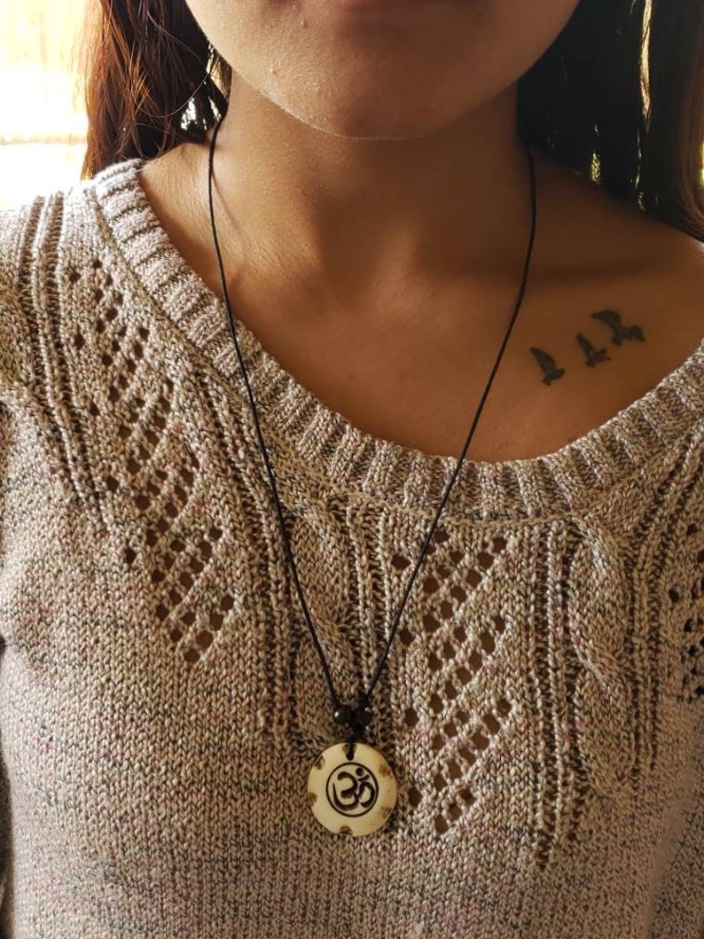 May include: A black cord necklace with a wooden pendant featuring the Om symbol. The pendant is carved with intricate details and has a light brown color.