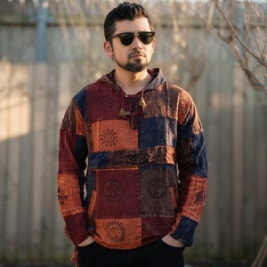 Puede incluir: Una sudadera con capucha y manga larga que presenta un diseño de parches en tonos naranja, rojo y azul oscuro. La tela está decorada con varios estampados circulares de soles y símbolos por toda la prenda, incluyendo un bolsillo tipo canguro.