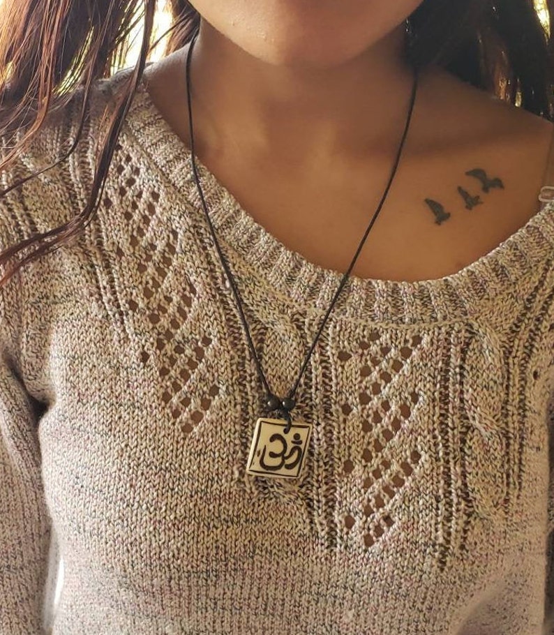 May include: A black cord necklace with a square pendant featuring the Om symbol in black and white. The pendant is made of ceramic and has a slightly textured surface.