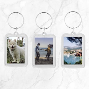 May include: Three clear plastic keyrings with silver rings. Each keyring has a different photo inside: a white dog, a couple on a beach, and a view of a town by the sea.