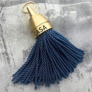Pode incluir: Um porta-chaves decorativo com borla, apresentando uma tampa de metal dourado gravada com a palavra CASA. A tampa está presa a um anel circular dourado e a um denso grupo de fios de corda torcidos azul-escuros que pendem em leque.