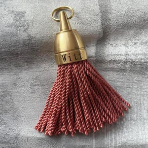 May include: A decorative tassel featuring a brushed gold metal cap with a circular ring at the top. The metal cap is engraved with the text "Witt" in a black sans-serif font. Attached to the cap is a dense cluster of dusty rose twisted cord.