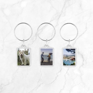 Op de afbeelding: Drie transparante sleutelhangers van plastic met zilveren ringen. Elke sleutelhanger heeft een rechthoekige foto-inzet. De foto-inzetten tonen een witte hond, een stel dat hand in hand loopt en een uitzicht op een haven.