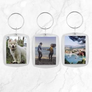 Op de afbeelding: Drie transparante sleutelhangers van plastic met zilveren sleutelringen. Elke sleutelhanger heeft een andere foto erin: een witte hond, een stel dat hand in hand op een strand staat en een schilderachtig uitzicht op een stad.