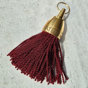 May include: A decorative tassel featuring a deep burgundy, multi-strand cord fringe attached to a brushed gold-tone metal cap. The metal cap is engraved with the text CHATEAU 4 and includes a circular metal ring at the top for hanging purposes.
