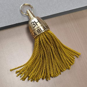 May include: A gold-toned metal tassel cap with a circular ring attachment, featuring the engraved text 3H and SA BLAN. The cap is connected to a dense, fan-shaped cluster of mustard yellow twisted cord fringe that hangs downward in a wide arc.