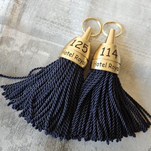 May include: Two navy blue tassel keychains with gold-toned metal caps. Each cap is engraved with the text Hotel Royal and a room number, 125 and 114 respectively. A circular gold metal ring is attached to the top of each conical cap.
