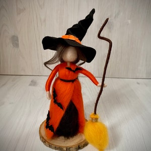 May include: A felt witch doll with long brown hair, wearing a black pointed hat with an orange band, an orange dress with black bats, and holding a brown stick with a yellow broom.