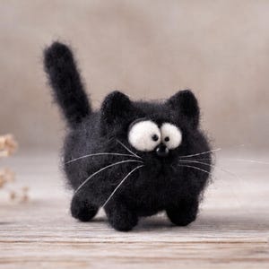 Può includere: Figurina di gatto in feltro nero fatta a mano con grandi occhi bianchi e lunghi baffi. Il gatto ha una coda soffice ed è su una superficie di legno. È un oggetto decorativo stravagante.