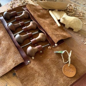 May include: A brown leather tool roll containing wood carving chisels with wooden handles. Also visible are wooden carvings of a bear and other tools, all on a wooden surface. The chisels are secured with leather straps.