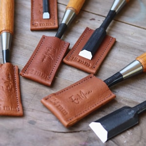 Puede incluir: Una colección de formones de madera con mangos de madera y hojas de metal, cada uno con una funda de cuero marrón. Las fundas están grabadas con medidas en milímetros, como 18 mm y 24 mm, y algunas tienen iniciales.