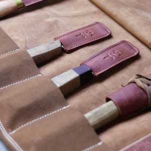May include: A brown leather roll with six slots for holding tools. The roll is open and shows three tools with brown leather sheaths. Each sheath has the text "Leather" embossed on it.