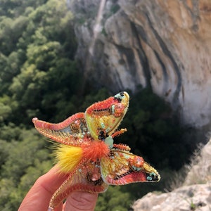 May include: A colorful butterfly brooch with yellow, orange, pink, and green sequins and beads. The brooch has a fluffy, yellow and orange feather accent.