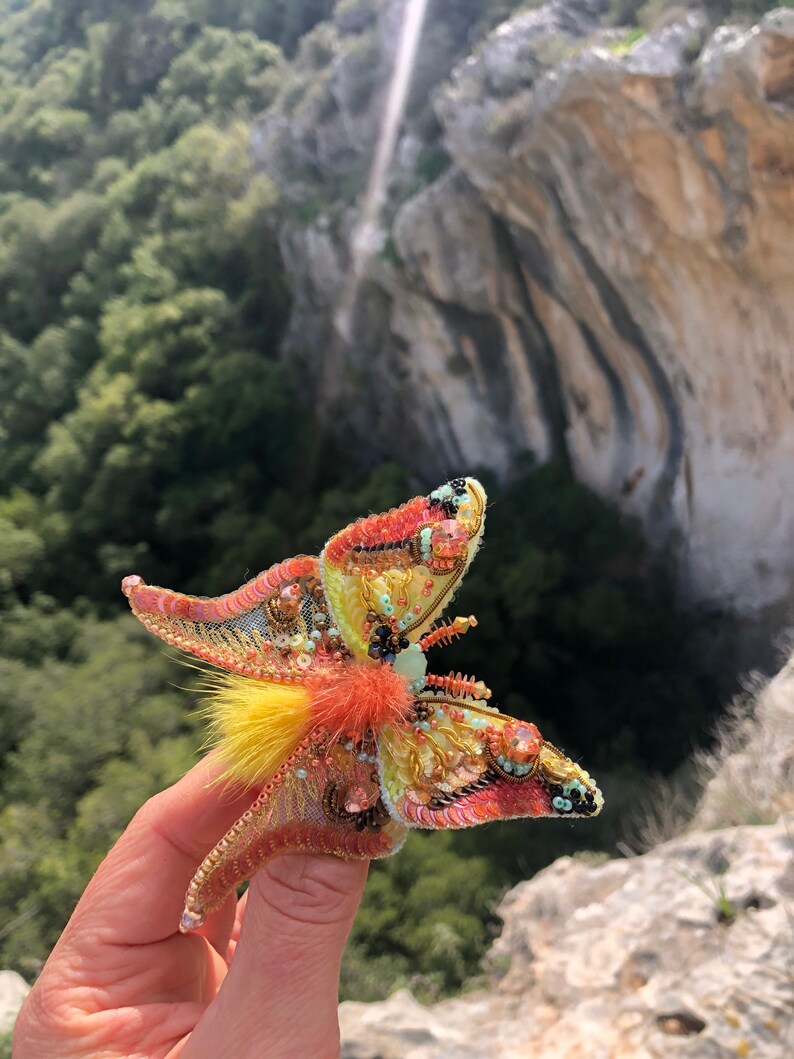 May include: A colorful butterfly brooch with yellow, orange, pink, and green sequins and beads. The brooch has a fluffy, yellow and orange feather accent.