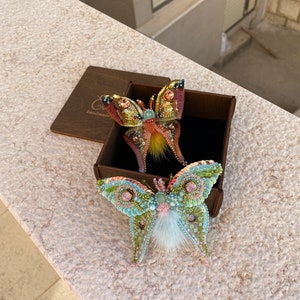 May include: Two beaded butterfly brooches in a brown box. One butterfly is yellow, orange, and pink with a fuzzy orange center. The other butterfly is green, blue, and pink with a white fuzzy center. The box has the word "handmade" on it.