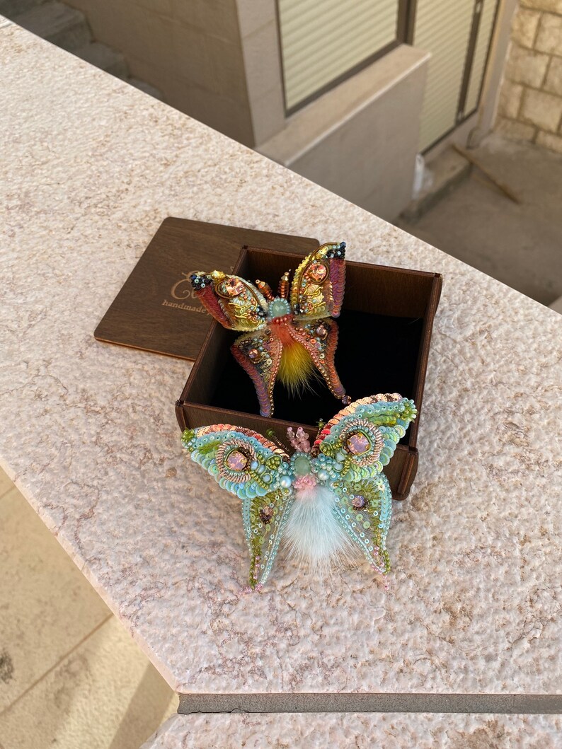 May include: Two beaded butterfly brooches in a brown box. One butterfly is yellow, orange, and pink with a fuzzy orange center. The other butterfly is green, blue, and pink with a white fuzzy center. The box has the word "handmade" on it.