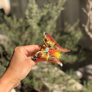 May include: A beaded butterfly brooch with red, orange, yellow, and green accents. The butterfly has a textured, iridescent finish and a small, light blue gem in the center.