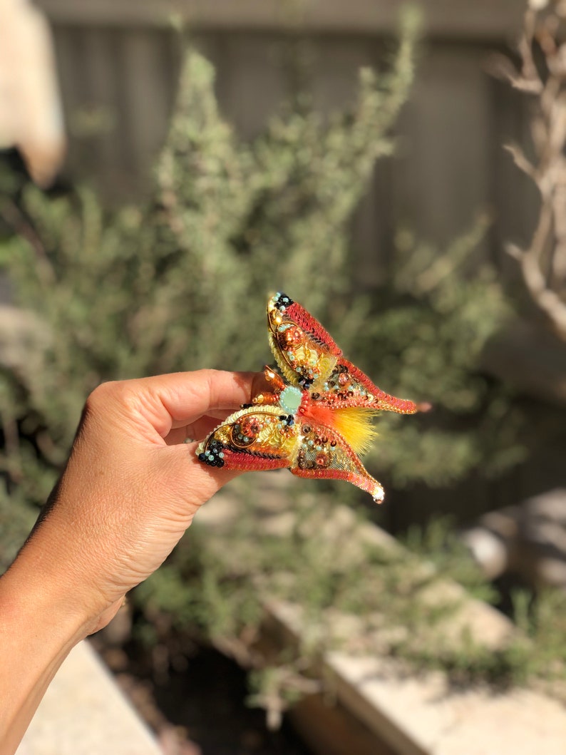May include: A beaded butterfly brooch with red, orange, yellow, and green accents. The butterfly has a textured, iridescent finish and a small, light blue gem in the center.