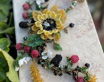 Könnte beinhalten: Eine Perlenkette mit einer Sonnenblume, Beeren und Blättern. Die Sonnenblume hat einen Kristallkern und die Beeren sind rot und schwarz.