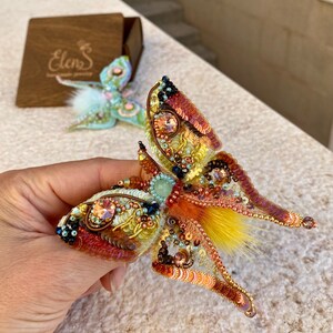 May include: A beaded butterfly brooch with iridescent wings, yellow and orange sequins, and a fluffy orange and yellow center. The brooch is handmade and features a delicate design.