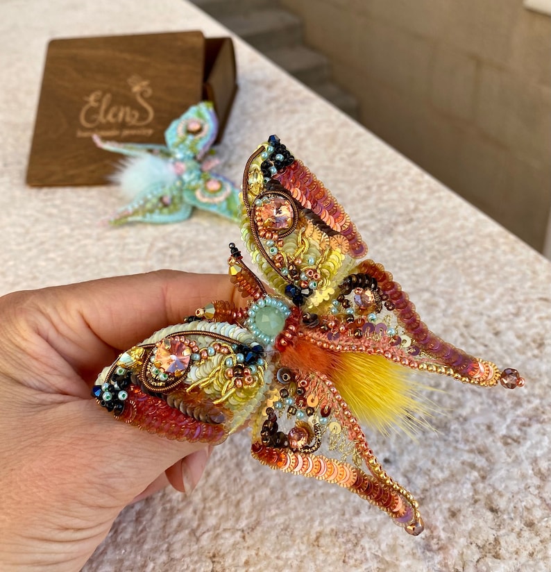 May include: A beaded butterfly brooch with iridescent wings, yellow and orange sequins, and a fluffy orange and yellow center. The brooch is handmade and features a delicate design.