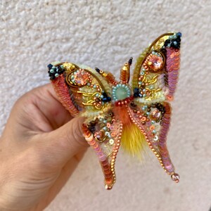 May include: A colorful butterfly brooch with iridescent sequins, beads, and feathers. The butterfly has a yellow, orange, pink, and green color scheme.