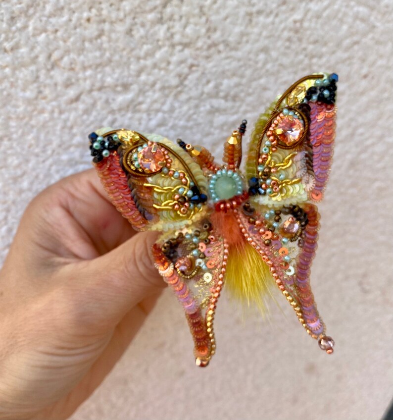 May include: A colorful butterfly brooch with iridescent sequins, beads, and feathers. The butterfly has a yellow, orange, pink, and green color scheme.
