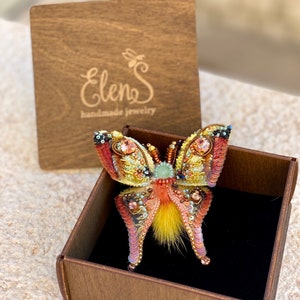 May include: A colorful butterfly brooch with yellow, orange, and pink sequins and beads. The brooch is in a wooden box with the text "Elens handmade jewelry" on the lid.