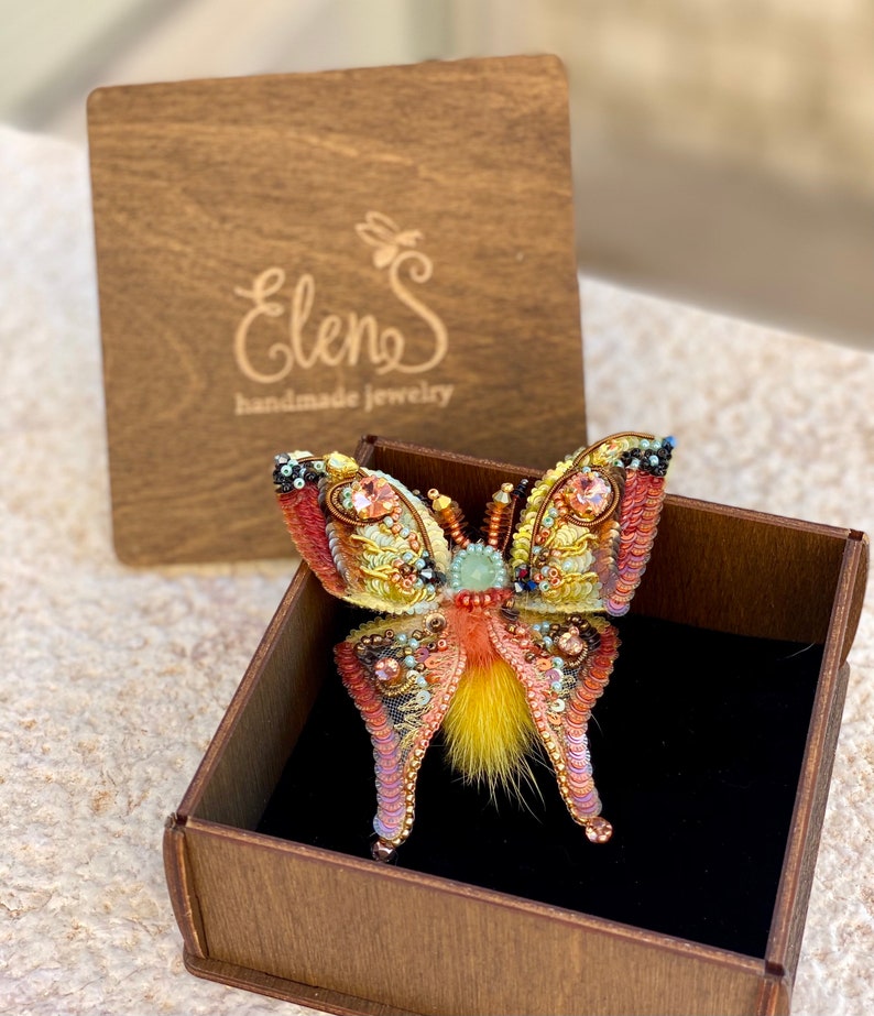 May include: A colorful butterfly brooch with yellow, orange, and pink sequins and beads. The brooch is in a wooden box with the text "Elens handmade jewelry" on the lid.