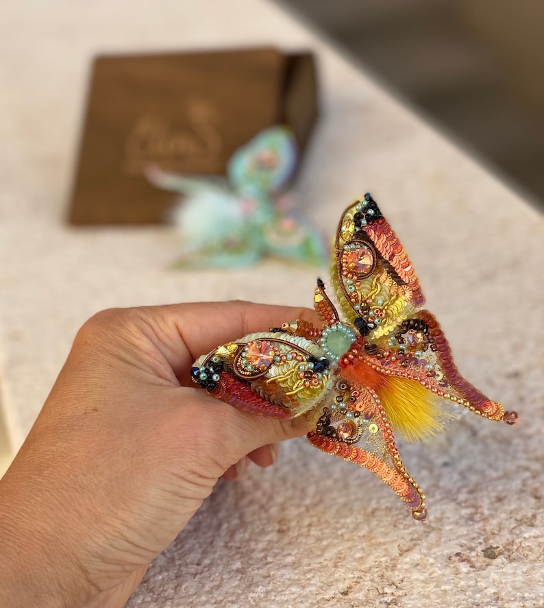 May include: A beaded butterfly brooch with orange, yellow, and green accents. The butterfly has a feathery texture and is made with iridescent beads.
