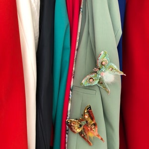 May include: A close-up of a row of clothing with a green blazer featuring two decorative butterfly brooches. One brooch is green and white, and the other is orange and gold.