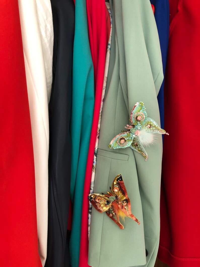 May include: A close-up of a row of clothing with a green blazer featuring two decorative butterfly brooches. One brooch is green and white, and the other is orange and gold.