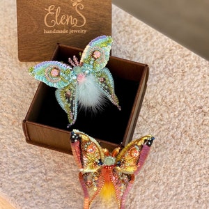 May include: Two handmade butterfly brooches in a wooden box. The top brooch is light blue and green with white fur accents. The bottom brooch is orange and pink with yellow fur accents. The box has the text "Elens handmade jewelry" on the top.