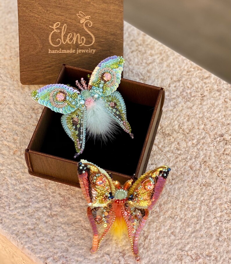 May include: Two handmade butterfly brooches in a wooden box. The top brooch is light blue and green with white fur accents. The bottom brooch is orange and pink with yellow fur accents. The box has the text "Elens handmade jewelry" on the top.