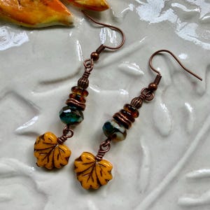 May include: A pair of dangle earrings featuring copper-toned ear wires, small amber and teal faceted glass beads, and a yellow ceramic maple leaf charm at the base. The earrings are constructed with copper wire wrapping and metal spacer beads.