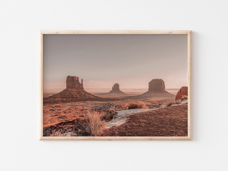 K&ouml;nnte beinhalten: Ein Panoramablick auf Monument Valley mit drei ikonischen Sandsteinfelsen, die aus der W&uuml;stenlandschaft aufragen. Der Himmel ist in einem sanften, blassen Rosa gehalten, und die Felsformationen sind in warmes, goldenes Licht getaucht.