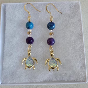 May include: Gold-coloured dangle earrings with a sea turtle design. Each earring has a gold hook, blue and purple beads, and a turtle pendant with a pearlescent shell. The earrings are displayed in a white box.