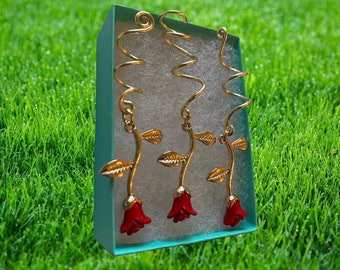 Puede incluir: Tres pendientes de rosa dorados con capullos de rosa rojos y hojas doradas se muestran en una caja color turquesa. Los pendientes tienen un diseño en espiral en la parte superior. El fondo es un césped verde borroso.
