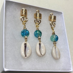 May include: Three gold-toned hair accessories with a spiral design at the top. Each accessory features a blue glass bead and a white seashell charm. The accessories are displayed in a white box.