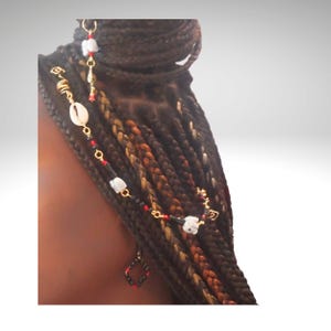 May include: A close-up of braided hair adorned with gold-toned hair jewelry. The jewelry includes a seashell, white beads, red and black beads, and a small, square, red and black charm. The braids are a mix of brown, black, and blonde.