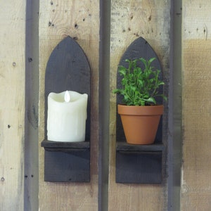 May include: Two wooden wall shelves with a gothic arch design. The left shelf has a white candle on it and the right shelf has a small potted plant with green leaves.