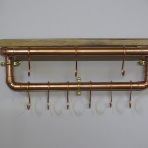 May include: Copper pipe shelf with a wooden top and hooks. The shelf is mounted on a white wall.
