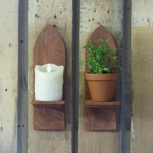 May include: Two wooden wall shelves with a pointed top. The left shelf holds a white candle and the right shelf holds a potted green plant.