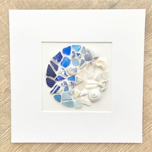 May include: A framed artwork featuring a circular arrangement of blue and white sea glass, ceramic shards, shells, and feathers. The composition transitions from dark to light blue, creating a gradient effect. The artwork is mounted on a white background.