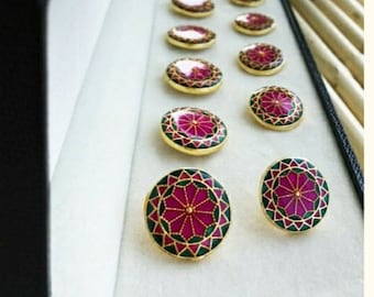 Puede incluir: Un juego de diez gemelos redondos de color dorado con un diseño geométrico rosa y verde. Los gemelos se muestran en una caja negra con un interior blanco.