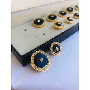 May include: A set of nine round, gold-toned buttons with black enamel centers and clear rhinestone accents. The buttons are arranged in a box.