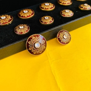 Może przedstawiać: Zestaw 12 ozdobnych guzików z pozłacanym rantem i czerwoną podstawą. Guziki mają biały kryształ górski w środku i złoty wzór kwiatowy.