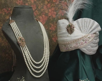 Puede incluir: Un elegante collar de perlas con un cierre decorativo se exhibe en un maniquí. Un turbante blanco con una pluma y una broche enjoyada se encuentra al lado. El fondo presenta un estampado floral y tela verde oscuro.
