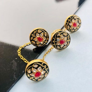 Puede incluir: Un conjunto de tres cierres de botón dorados con esmalte negro y piedras preciosas rojas y blancas. Los botones están conectados por una cadena dorada.