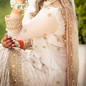 May include: A white bridal outfit with intricate gold embroidery and a matching veil. The outfit features a long-sleeved top and a flowing skirt.
